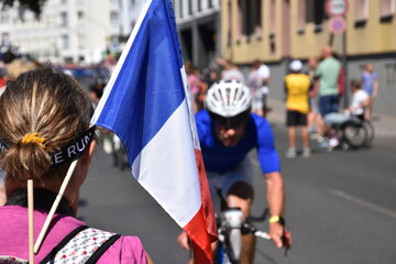 Eine französische Fahne eines Fans im Mittelpunkt des Bildes. Im Hintergrund ist ein Triathlet auf...