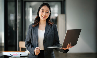 Beautiful asian businesswoman holding laptop computer standing in office, smiling at camera.