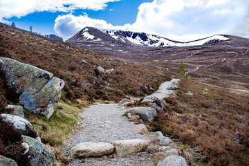 Hiking trail, Cairngorms National Park, Scottish Highlands, Scotland, popular hiking and skiing area. A tall snow capped peak in distance, blue sky with clouds. Curved trail, rocks in foreground.
