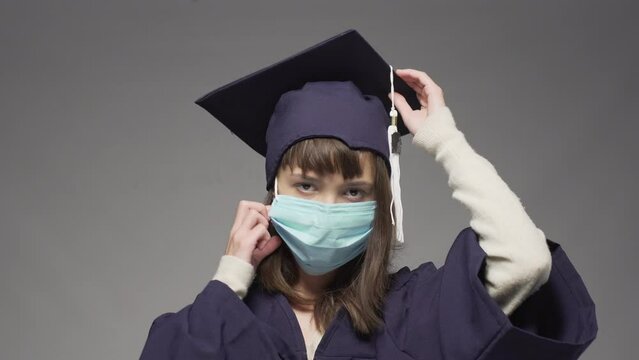 Graduation girl wearing cap and taking off  protective face mask. Female student smiling against gray background.