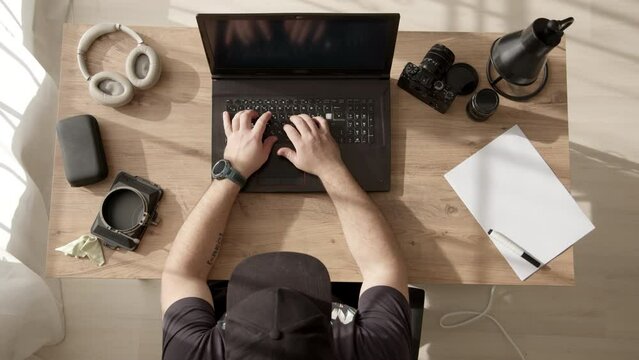 Unrecognizable male freelancer in black cap is sitting at table and typing on laptop at home. Guy on online training in office. Programmer at work at computer, top down view.