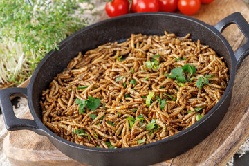 Cooked fried edible mealworms with spice and herbs in frying pan on wooden board. Meal worms as...