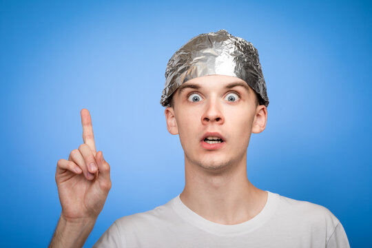 Paranoid young man wearing tin foil hat pointing up studio shot on blue background
