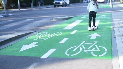 Girl on a kick scooter rides on green bike path in a city. child on scooter riding in the bike lane. white Bicycle sign on green road traffic laws, crosswalk and cars.
