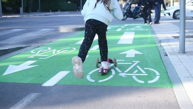 Girl on a kick scooter rides on green bike path in a city. child on scooter riding in the bike lane. white Bicycle sign on green road traffic laws, crosswalk and cars.