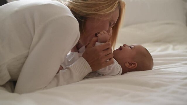 Smiling baby and mother in white t-shirts play on bed. Side view. Mother kiss and laugh with baby. love and care in happy family. First year of life and knowledge of world around.
