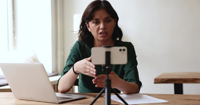 Engaged Asian blogger woman speaking at webcam on mobile phone, working, giving webinar, shooting video, recording podcast, making conference call to client from co-working office space