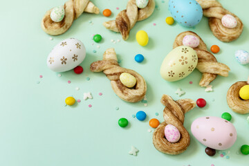 Fototapeta na wymiar Colors Easter eggs with Easter rabbit-shaped buns puff pastry with cinnamon on pastel green background. Happy Easter Holidays.