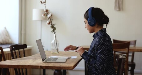 Confident young Indian blogger woman in wireless headphones recording podcast, using laptop for online communication, making video call, waving hello, bye hand, speaking, listening, writing notes