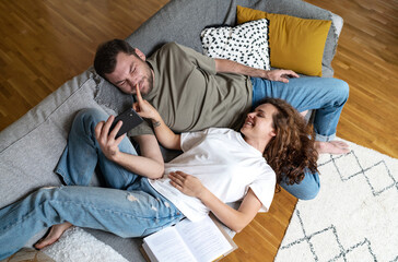 Playful couple lying down on sofa at home man using smart phone girlfriend touching boyfriends nose...