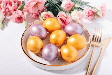 Gold and purple painted Easter eggs in pastel colors in a plate on a white cotton tablecloth