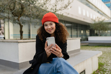 pretty curly woman in city street in autumn coat, using smart phone, red knitted hat
