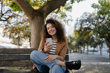 pretty curly smiling woman walking in city street in stylish jacket, using smartphone