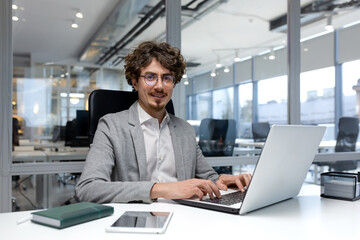 Portrait of hispanic businessman, man with curly hair and beard wearing glasses smiling and looking at camera, programmer typing on laptop inside bright office.