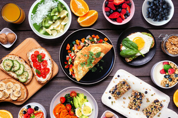Healthy breakfast food table scene. Flat lay view on a dark wood background. Omelette, nutritious bowl, toasts, granola bars, smoothie bowl, yogurts and fruits.