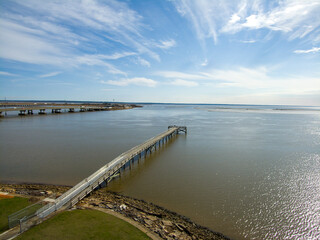 A long brown wooden pier of the rippling waters of Mobile Bay with cars driving on the highway and...