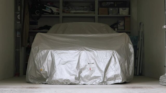 A sports car is parked under an awning in a bright home garage. Footage. Opens the gates of a white garage, inside a car closed with a cloth and shelves with a tool in the background