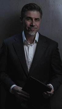 Portrait of happy mature businessman with tablet computer. Entrepreneur standing in front of dark loft wall. Older, middle aged, mid adult, man in his 40s or 50s in shirt and jacket, business casual.