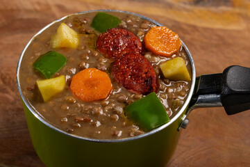 Stewed lentils with chorizo, apple or potato, green pepper and carrot in a green metal saucepan and on a wooden board. Concept of traditional and rustic cooking.