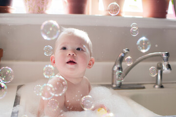 Baby taking bath in the kitchen sink. Child playing with foam and soap bubbles in the sunny bathroom with a window. Water fun for kids. Hygiene and skincare for children.