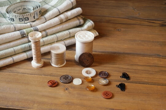 Layers of Green and Cream Plaid Fabrics, Spools of Thread, Buttons and Tape Masure on Medium Brown Panelsi