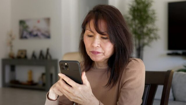 Happy attractive middle age woman relaxing excited successful shopping online on smartphone credit card sitting at table in casual leisure home 