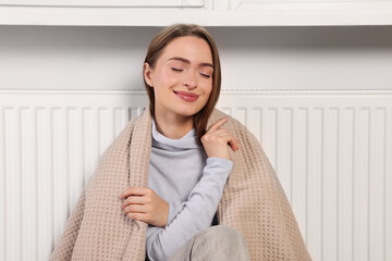 Woman with blanket sitting near heating radiator indoors