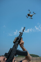 A man aims to shoot a rifle at a flying drone against a blue sky. 