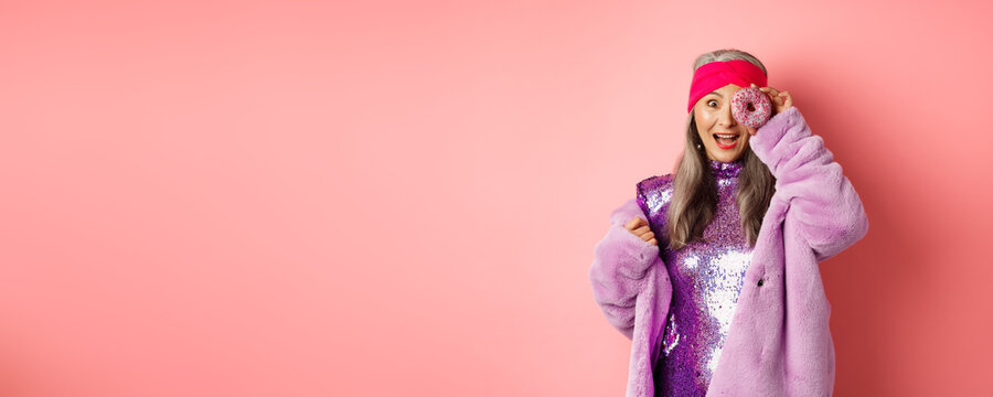 Food And Fashion Concept. Beautiful Asian Senior Woman In Disco Dress And Faux Fur Coat Looking Through Donut Hole With Amazed Face, Standing Happy Over Pink Background