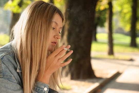 Young woman smoking cigarette in park on sunny day, space for text