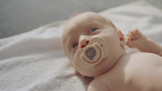 Adorable cute naked toddler sucking pacifier relaxing and lookng around while lying on bed, feeling calmness. Close-up and slow-motion shot