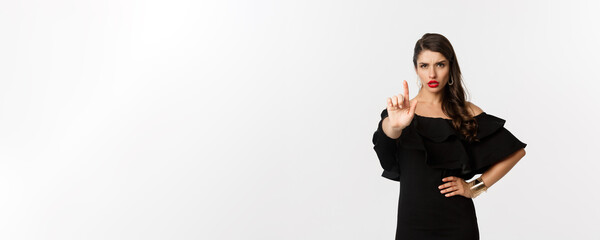 Fashion and beauty. Confident and serious lady in black dress, showing finger in stop gesture,...