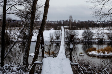 An old suspension wooden bridge over an unfrozen river on a frosty winter day.