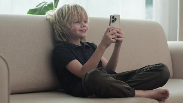 Happy smiling blonde boy using looking phone and sitting home on sofa, holding smartphone two hands.