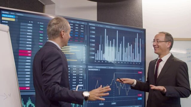 Cheerful colleagues analysing graphics on big digital screen. Data engineers pointing at monitor with pen, talking, analysing financial statistics in meeting room. Teamwork, business concept