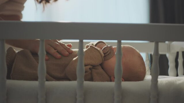 A cute baby with a pacifier in his mouth, lying in a baby crib on a white blanket. Mother gently touching baby's belly in slow-motion