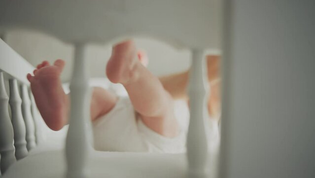 Close-up view of the baby moving his legs around while lying in the crib at morning. Mother gently stroking baby in slow-motion