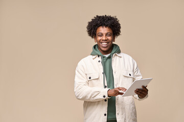 Happy cool curly African American teenager student boy wearing white jacket holding pad using digital tablet tab computer technology for online education standing isolated on beige background.