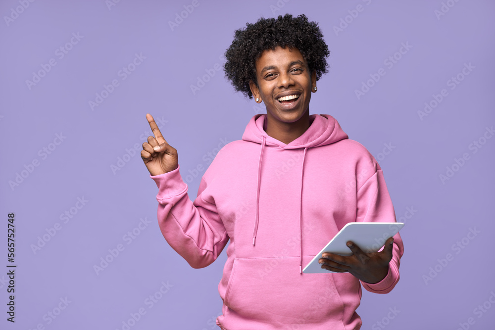 Wall mural Happy smiling African American teen boy student wearing pink hoodie holding using digital tablet pointing finger aside at copy space advertising, presenting isolated on light purple background. - Wall murals
