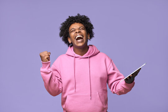 Happy euphoric excited African teen winner student boy holding digital tablet computer screaming winning prize in online game, celebrating great result with yes gesture isolated on purple background.