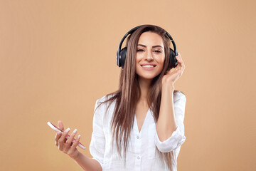 Portrait of smiling brunette woman in headphones with smartphone listening music over beige background. Girl uses wireless earphones.