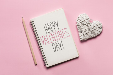 Above view flat lay composition of sketchbook with hand written St. Valentines day greetings, pencil and decorative rattan heart on pastel pink background