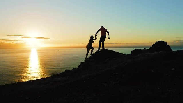 Couple of travelers climb the mountain during sunset and look at ocean sunset. Man gives a helping hand to womn. Travel vacation adventure teamwork concept.