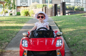 Caucasian baby in a white hat riding in an electric convertible red sports car. child driving a car on a summer day	