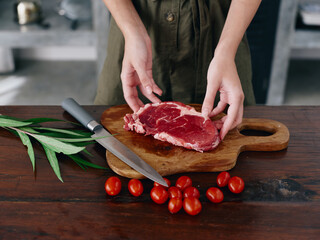 Woman with knife in hand cutting fresh steak meat for roasting in kitchen with salt pepper and other spices on table, red cherry tomatoes and herbs, preparing dinner. wooden table, top view.
