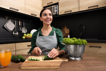 Woman in apron cutting cucumber at wooden table indoors