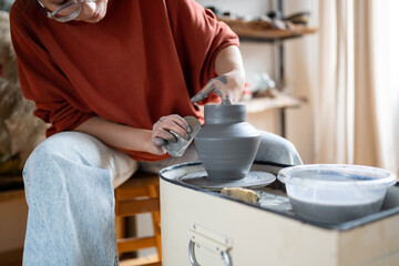 Attentive woman sits in pottery workshop and diligently does what she loves, which brings income and pleasure. Female makes aesthetic items for home with own hands. Working with clay for mental health