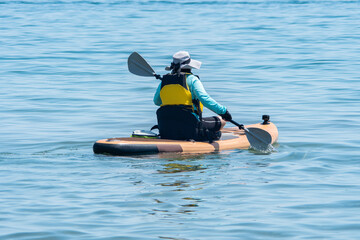 Woman paddle on board at calm lake water during warm summer. Stand up paddle board with special seating chair, wet suit and life jacket for paddling. Paddle SUP board sport and active recreation.