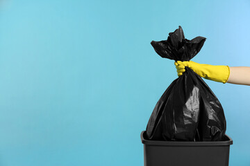 Janitor in rubber glove holding trash bag full of garbage over bucket on light blue background, closeup. Space for text - obrazy, fototapety, plakaty