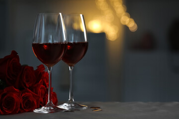 Glasses of red wine and rose flowers on grey table against blurred lights, space for text. Romantic atmosphere
